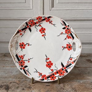 May include: A round white ceramic serving tray with two integrated side handles. The surface features a decorative pattern of bright red flowers on black branches, accented by geometric black and red grid lines and gold-toned trim on the edges.