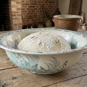 May include: A large, antique white ceramic bowl with a blue floral design holds a loaf of bread. The bowl sits on a rustic wooden table, with a brick fireplace and other kitchenware in the background.