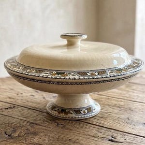 Peut inclure: Plat de service crème avec couvercle et piedestal. Le plat présente une bordure décorative avec des motifs géométriques et floraux verts, dorés et noirs. Le couvercle a une petite poignée arrondie.