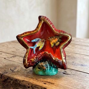 Peut inclure: Un plat en céramique en forme d'étoile avec un intérieur rouge vif et une bordure dorée. Deux poissons colorés nagent à l'intérieur. La base est verte et le plat repose sur une surface en bois. Une pièce décorative.