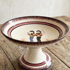 Peut inclure: Un compotier sur pied de couleur crème avec un large bord orné d'un motif répétitif marron foncé et rose. L'intérieur du bol présente une scène peinte de deux personnages. La base est crème avec une bande marron foncé et rose.
