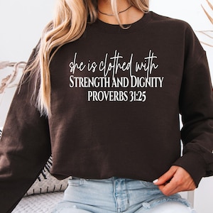 May include: A dark brown sweatshirt with the text "she is clothed with STRENGTH AND DIGNITY PROVERBS 31:25" in white script. The sweatshirt is long-sleeved and has a crew neck. The image is of a person wearing the sweatshirt.