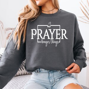 Peut inclure: Sweat-shirt gris foncé avec l'inscription blanche "PRAYER changes things". Le mot "PRAYER" est en gras, en lettres majuscules, et "changes things" est en écriture cursive. Un graphisme de mains jointes est au-dessus du mot "PRAYER".