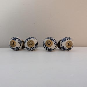 May include: Four ceramic cabinet knobs with a white base, adorned with blue floral patterns and gold-colored metal hardware. The knobs are arranged in a row, showcasing their decorative design.