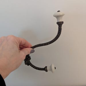 May include: A decorative wall hook with a dark brown, twisted metal design. The hook features two white ceramic knobs with a black dot on top. The hook is held by a hand against a white wall.