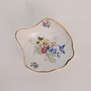 May include: A white porcelain trinket dish with an irregular, organic shape and a gold-painted rim. The center features a floral bouquet with yellow, pink, and blue flowers, green foliage, and a small blue butterfly. Smaller floral accents appear.
