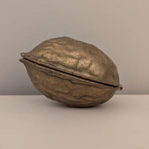 May include: A bronze-colored, walnut-shaped container with a hinged lid. The container has a textured surface and a small, round clasp. The object is resting on a white surface.