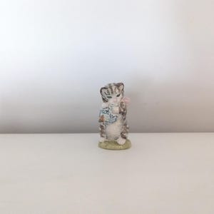 May include: A small ceramic figurine of a grey and white tabby kitten standing on a light green base. The kitten is holding a blue and white checkered ball of yarn with a small pink accent visible near its head, rendered in a detailed style.