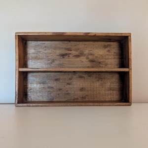 May include: A rectangular, two-tiered wooden shelf with a natural, aged finish. The shelf has a simple design with a horizontal divider creating two compartments. The wood grain is visible, adding to its rustic charm.