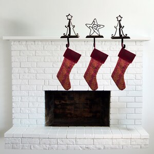 May include: Three Christmas stockings with deep red velvet cuffs and gold-patterned fabric bodies, each hanging from a black metal stocking holder. Two holders feature a star-topped tree design, while the center holder features a star design.
