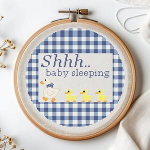 May include: A cross-stitch embroidery hoop with a blue and white gingham design. The text "Shhh.. baby sleeping" is stitched above a duck and three ducklings. The embroidery is set in a wooden hoop, ideal for a nursery.