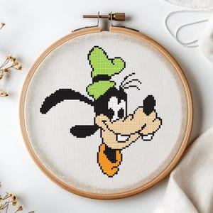 May include: A cross-stitch embroidery hoop featuring a pixelated image of Goofy, the Disney cartoon character. The design includes Goofy's face with a green hat, black ears, and an orange collar. The embroidery is set in a wooden hoop.