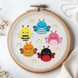 Op de afbeelding: Een borduurring met kruissteek met vijf kleurrijke insectenontwerpen: een gele bij, een blauw insect, een roze insect, een oranje insect en een rode lieveheersbeestje. Elk insect heeft vleugels en is omgeven door kleine regenbogen.
