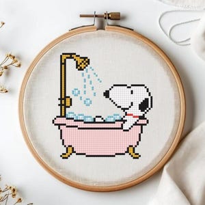 May include: Cross-stitch embroidery in a wooden hoop features a cartoon dog in a pink bathtub with bubbles and a shower head. The dog is white with black ears and a red collar. The background is white.