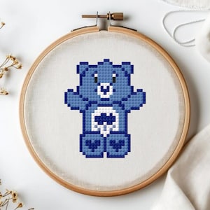 May include: A cross-stitch design of a blue bear with a white belly and heart-shaped design, displayed in a wooden embroidery hoop. The bear is made of pixelated squares, and the hoop is set against a white background with floral accents.