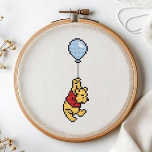 Könnte beinhalten: Kreuzstich-Stickerei eines gelben Bären, der einen blauen Ballon hält. Der Bär trägt ein rotes Hemd. Die Stickerei befindet sich in einem Holzreifen auf weißem Hintergrund.