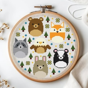 Op de afbeelding: Een borduurring met kruissteek met een bosdierenontwerp. Het ontwerp bevat een beer, vos, wasbeer, adelaar, konijn en das, samen met bomen en bloemaccenten. De dieren zijn in tinten bruin, oranje, grijs en zwart.