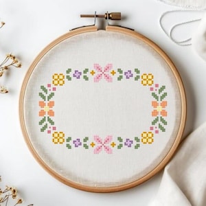 Puede incluir: Un bastidor de bordado de punto de cruz con un diseño floral en una variedad de colores. El diseño incluye flores rosas, amarillas, naranjas, verdes y moradas dispuestas en forma ovalada. El bastidor es de madera.