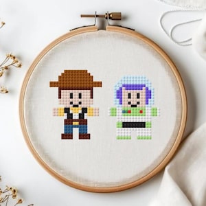 May include: A cross-stitch embroidery hoop featuring pixelated images of Woody and Buzz Lightyear from Toy Story. Woody is in a brown hat and Buzz is in a space suit. The hoop is set against a white background.