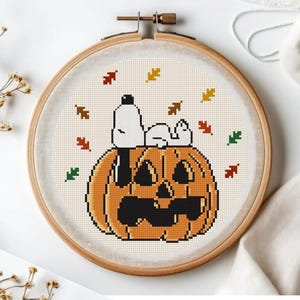 May include: A circular wooden embroidery hoop containing a cross-stitch pattern of a white dog resting on top of a large orange jack-o'-lantern. The design is surrounded by small, scattered autumn leaves in shades of red, yellow, brown, and green.