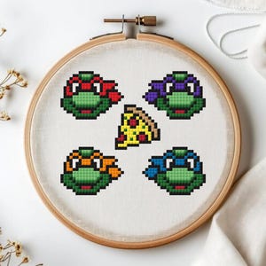 Puede incluir: Bordado de punto de cruz de las Tortugas Ninja, cada una con una máscara de diferente color: rojo, morado, naranja y azul. También se incluye una porción de pizza en el diseño. El bordado se muestra en un aro de madera.