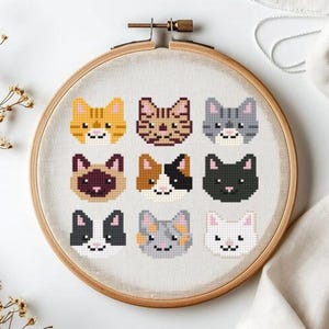 May include: A round embroidery hoop with a canvas featuring nine pixelated cat faces in various colors and patterns. The cats include orange tabby, brown tabby, gray, Siamese, calico, black, tuxedo, gray and orange, and white cats. The hoop is made of wood.