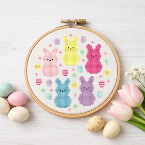 May include: A cross-stitch embroidery hoop featuring five colourful bunny figures in pink, yellow, and blue. The design includes pastel-coloured eggs and decorative elements, perfect for Easter. The wooden hoop is secured with a brass clasp.