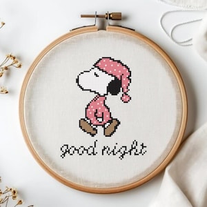 Könnte beinhalten: Kreuzstich-Stickerei in einem Holzrahmen mit Snoopy in rosa Schlafanzug und Nachtmütze. Die Worte "good night" sind darunter gestickt. Die Stickerei befindet sich vor einem weißen Hintergrund.