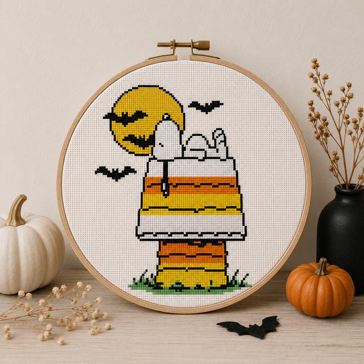 May include: A circular wooden embroidery hoop containing a cross-stitch pattern of a white dog lying on a candy corn colored doghouse. A large yellow moon and black bats are in the background. The design features horizontal stripes of orange, yellow, and white.