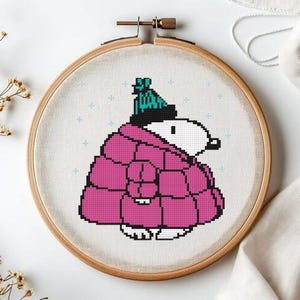 May include: Cross-stitch embroidery in a wooden hoop features a cartoon character wearing a pink quilted coat and a teal and black hat. The character has a white face and black features. Light blue cross stitches are scattered around the character.