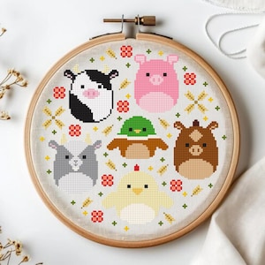 Op de afbeelding: Een borduurring met kruissteek met een ontwerp van boerderijdieren. Het ontwerp omvat een koe, een varken, een eend, een paard, een geit en een kip. De dieren zijn in een pixelachtige stijl, met een lichtbeige achtergrond en een houten ring.