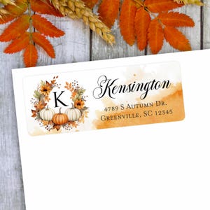 May include: A white rectangular label with fall-themed graphics. The label features a wreath of sunflowers and pumpkins, with the letter "K" in the center. The word "Kensington" is written in elegant script, along with an address.