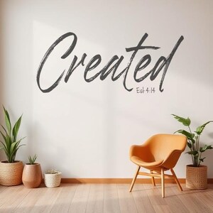 May include: A wall decal with the word "Created" in a stylized font, with the text "Est 4:14" below it. The decal is on a white wall, with a brown chair and plants in the foreground.