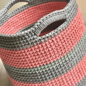 May include: A crocheted storage basket with two handles. The basket is made with pink and gray yarn in a striped pattern.