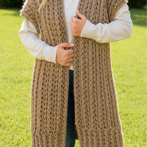 May include: A long, open-front, sleeveless cardigan in a warm brown color. The chunky knit design features a textured pattern. The cardigan is worn over a white long-sleeved shirt and blue jeans. The garment is knee-length.