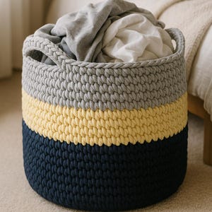 May include: A gray, yellow, and navy blue crocheted storage basket with a handle. The basket is filled with clothes.