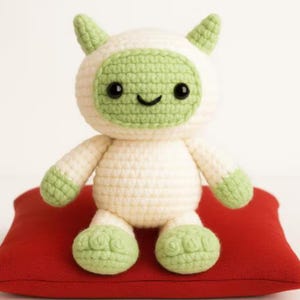 May include: A handmade crochet doll in the shape of a friendly creature. The doll has a white body with green accents on the face, feet, and horns. It sits on a red pillow. The doll has black eyes and a smiling mouth.