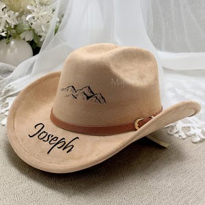 May include: A tan cowboy hat with a brown leather band and gold buckle. The hat features a black mountain range graphic and the name "Joseph" in script. The hat has a wide brim and a classic cowboy hat shape.