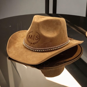 May include: A brown suede cowboy hat with a gold beaded trim and a leather band with the initials "M/S" engraved in a circle.