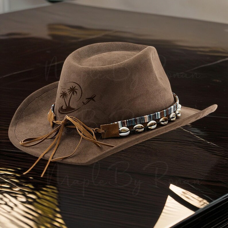 Op de afbeelding: Een bruine cowboyhoed met een leren band en schelpaccenten. De hoed heeft een ontwerp met palmbomen en een vliegtuig op de kroon.