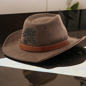 May include: A brown cowboy hat with a decorative leather band and a black rose and skeleton hand design. The hat has a wide brim and a classic cowboy hat shape. The hat is made of a soft material.