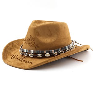 Op de afbeelding: Een bruine su&egrave;de cowboyhoed met een decoratieve band van schelpen en een bloemenmotief. De hoed is gepersonaliseerd met de naam "William".