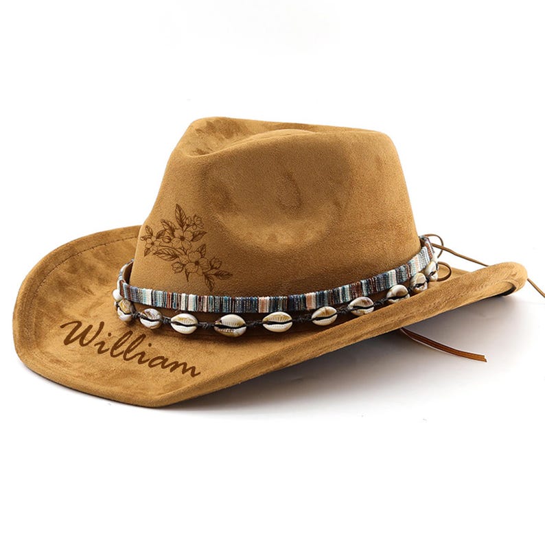 Op de afbeelding: Een bruine su&egrave;de cowboyhoed met een decoratieve band van schelpen en een bloemenmotief. De hoed is gepersonaliseerd met de naam "William".