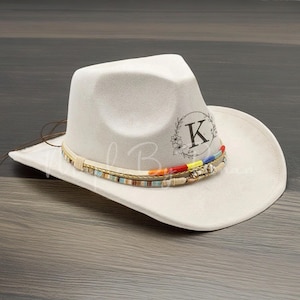 May include: A white cowboy hat with a brown leather band and colorful beads. The hat has a floral design with the letter "K" in the center.