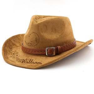 May include: A tan cowboy hat with a brown leather band and buckle. The hat features floral and crescent moon designs, along with the name "William" engraved on the brim. The hat is made of a soft, suede-like material.