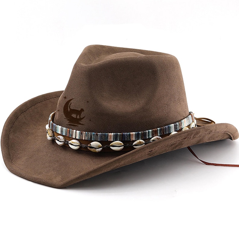Op de afbeelding: Bruine su&egrave;de cowboyhoed met een decoratieve band van schelpen en een maan- en kattenontwerp.