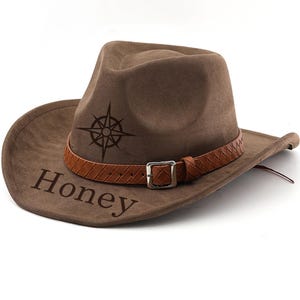 May include: A brown cowboy hat with a compass rose design and the word "Honey" printed on the brim. The hat features a brown leather band with a silver buckle. The hat is made of a soft, suede-like material.