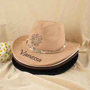 May include: A stack of cowboy hats in various colors, including tan, white, brown, and black. The top hat is tan with a decorative band and the name "Vanessa" printed on the brim. An illustration of a ram's skull and roses is on the crown.