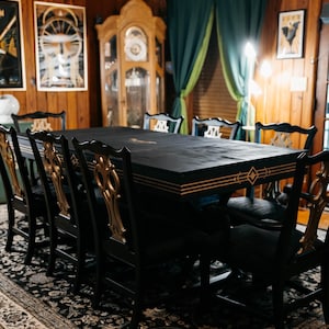 May include: A black dining table with gold accents and matching chairs. The chairs have ornate gold detailing on the backs. The table is set on a patterned rug in a room with wood paneling and artwork.