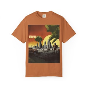May include: Orange t-shirt featuring a retro-style illustration of a post-apocalyptic cityscape with a robot, a vintage car, and the text "ATOMIC AGE". The artwork is set against a red and yellow sky with dark green smoke.